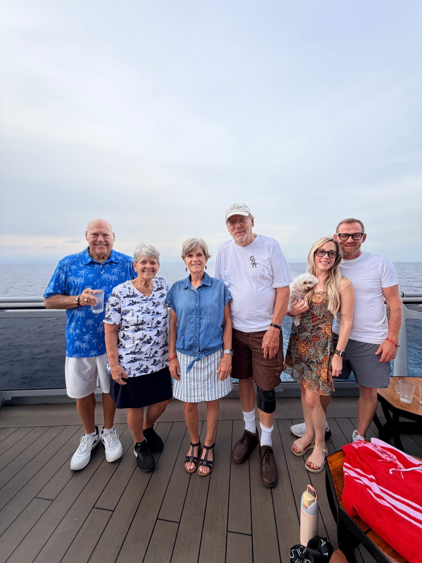 Six smiling guests, one holding a small white dog, on Scarlet Lady's open deck at sea