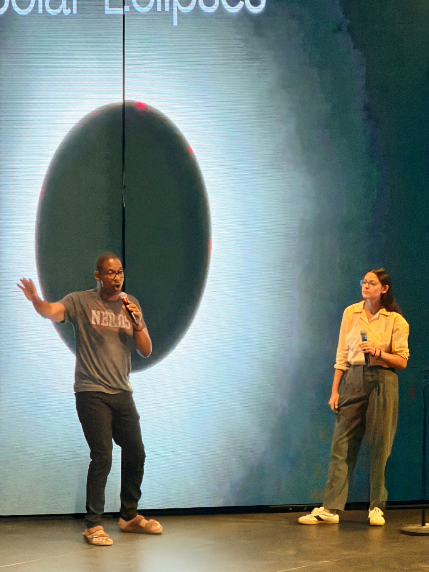 Two presenters on stage in front of a large solar eclipse graphic during Scarlet Lady's eclipse photography briefing