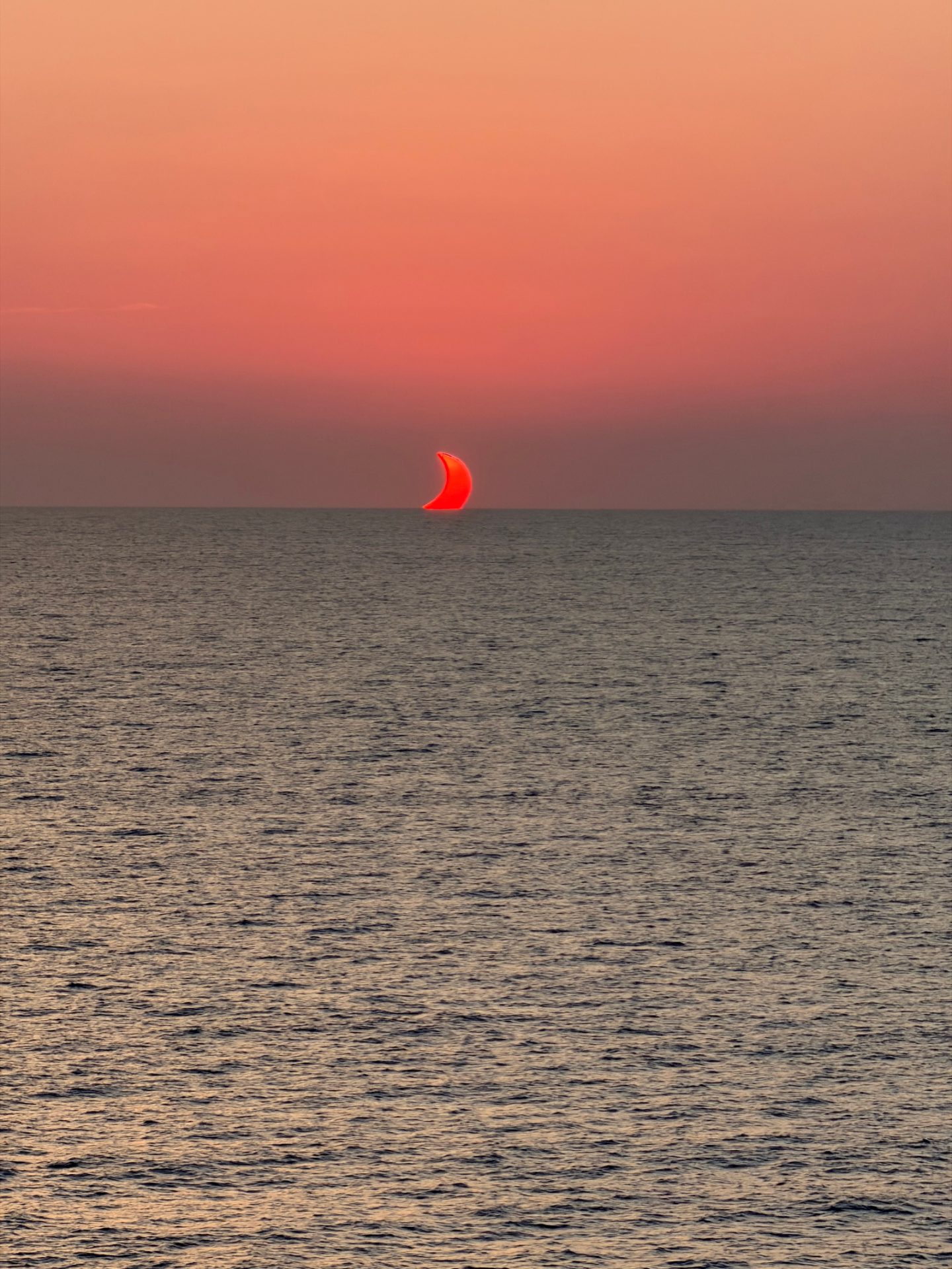 A red crescent sun partially eclipsed low over the Mediterranean horizon, seen from the sea